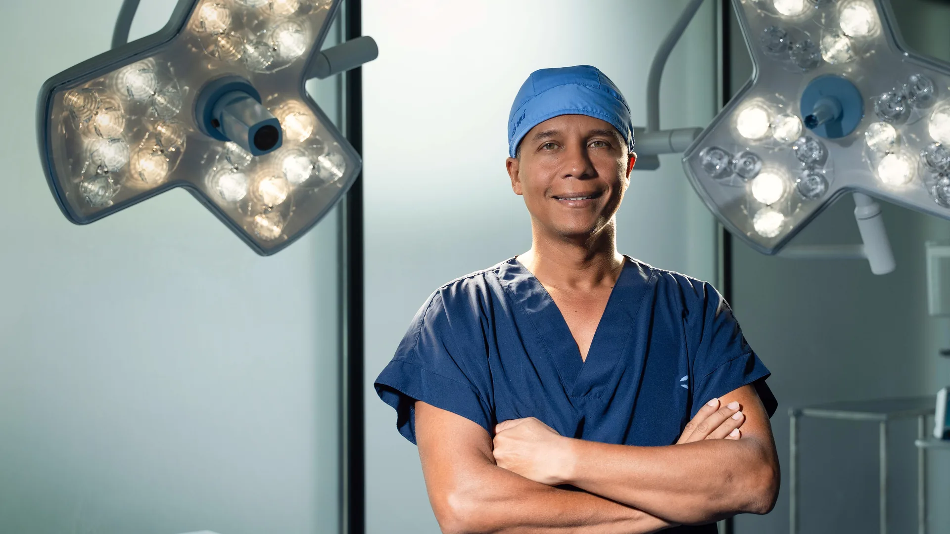 Dr. Fulvio Correa, plastic surgeon in Cartagena, Colombia, in the operating room wearing navy scrubs, smiling with arms crossed
